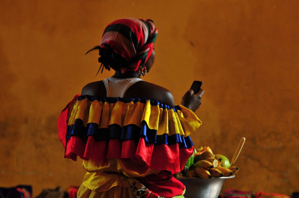A lady selling fruit in a colourful traditional dress using a mobile phone in a tropical city of Cartagena, Colombia.